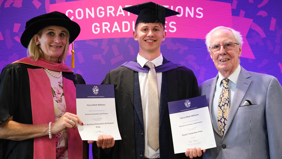 Three people at a graduation ceremony; two in academic regalia holding certificates, one in formal attire.